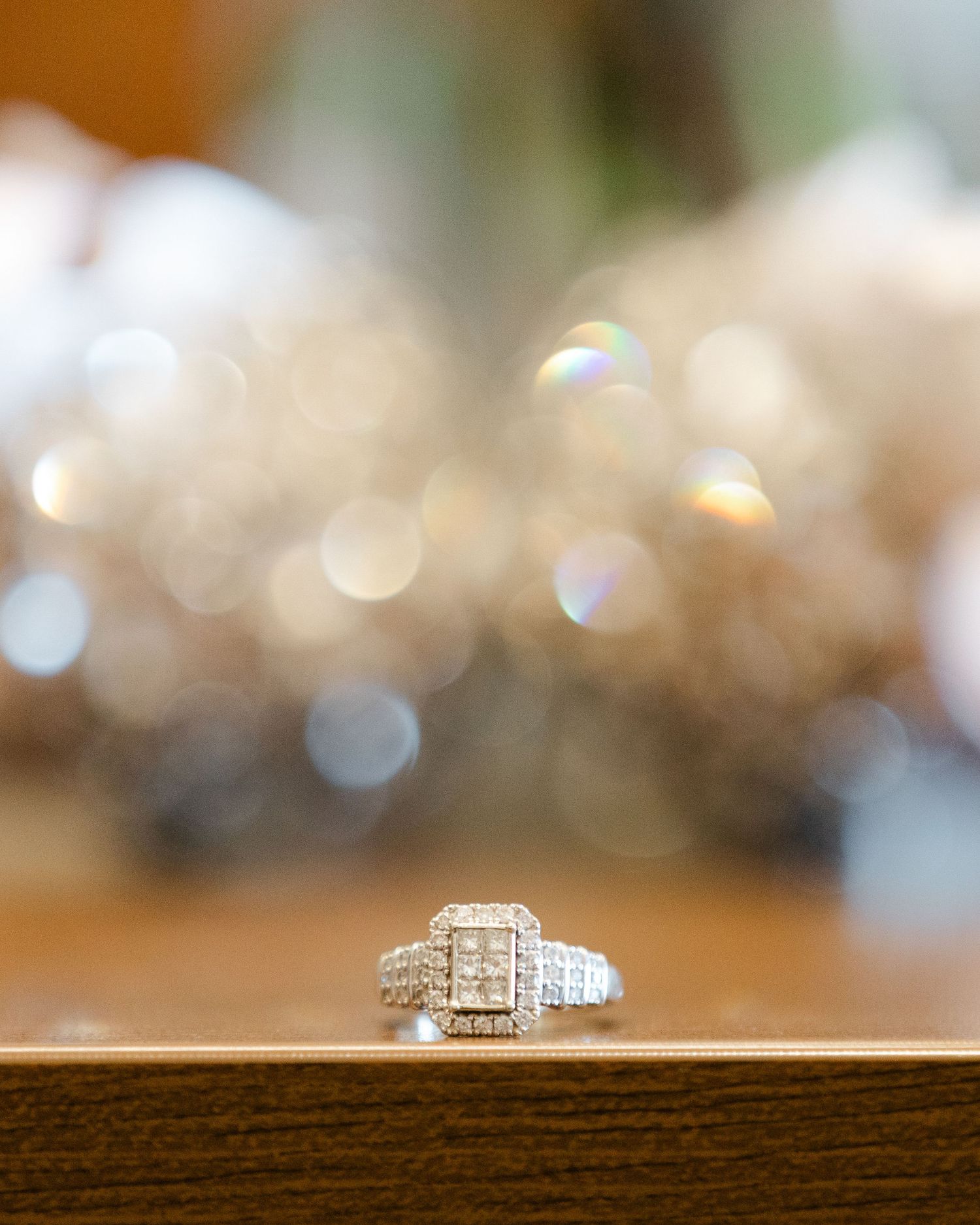 Elegant diamond engagement ring with sparkling stones displayed on dark wooden surface with soft bokeh background.