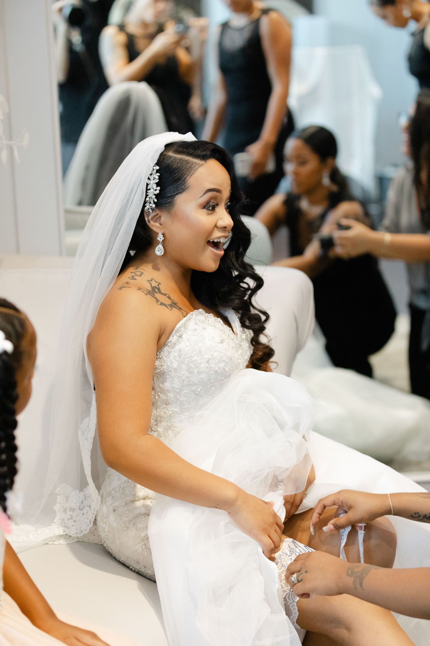 Bride in a white lace wedding dress sits gracefully while having her garter adjusted during the wedding reception.
