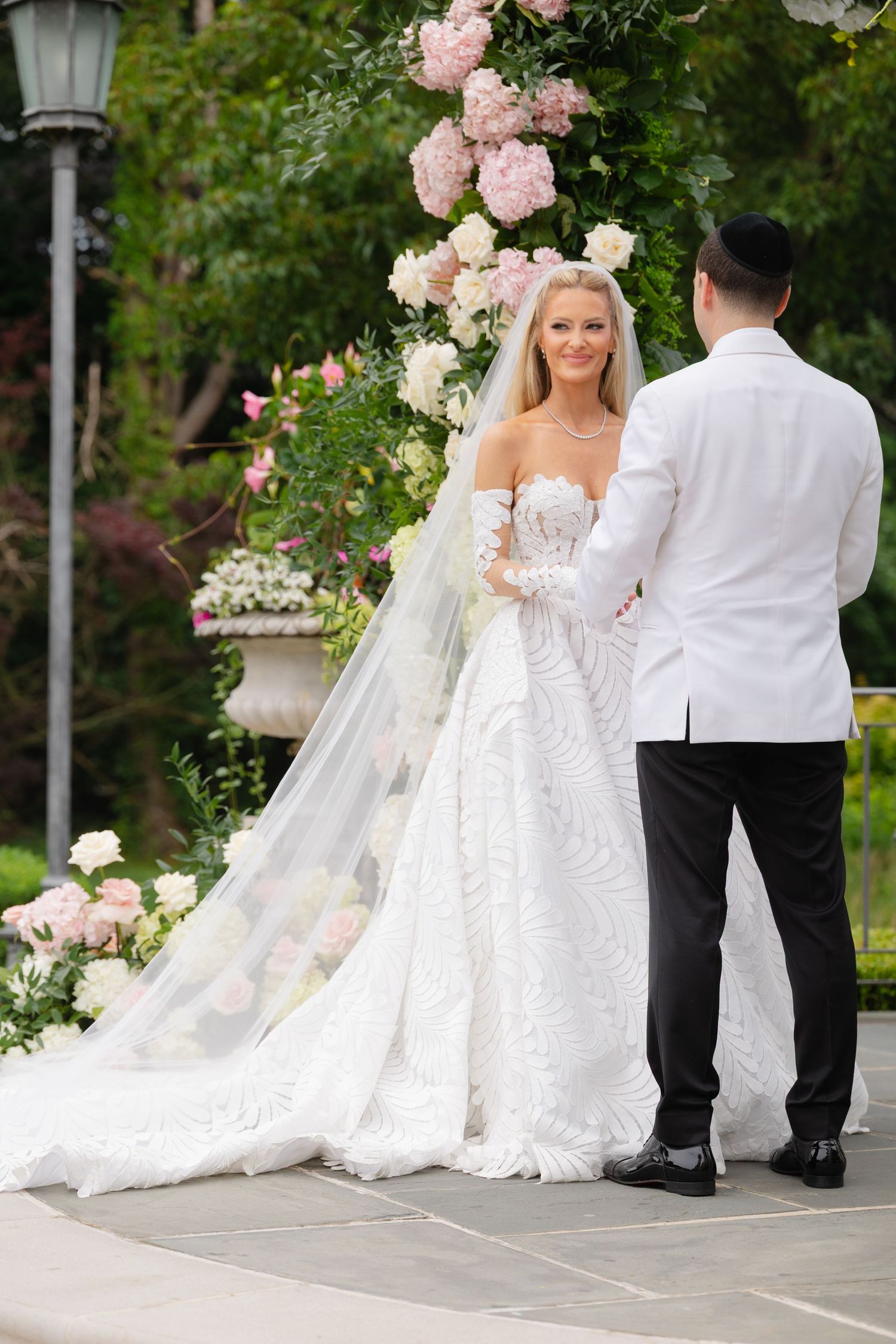 A bride and groom share romantic moments in a garden setting with pink roses and white floral arrangements.
