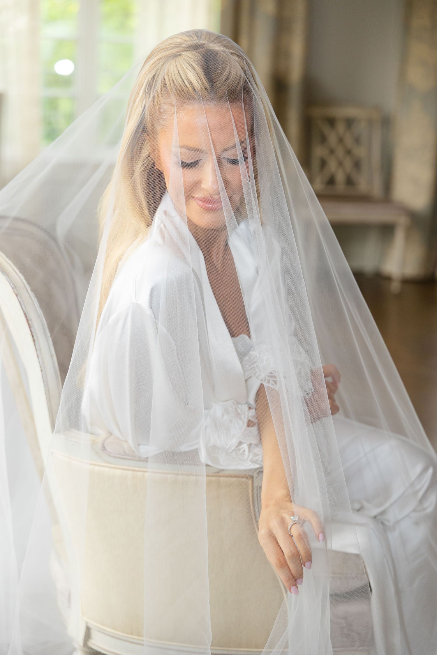 A bride in a white robe sits elegantly on a chair while wearing a flowing veil during wedding preparations.