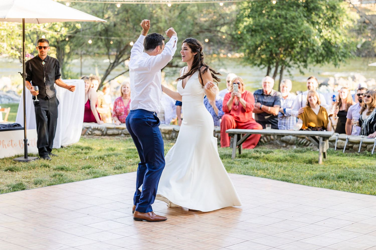 A couple dances together in an outdoor setting surrounded by guests and trees during a wedding celebration.