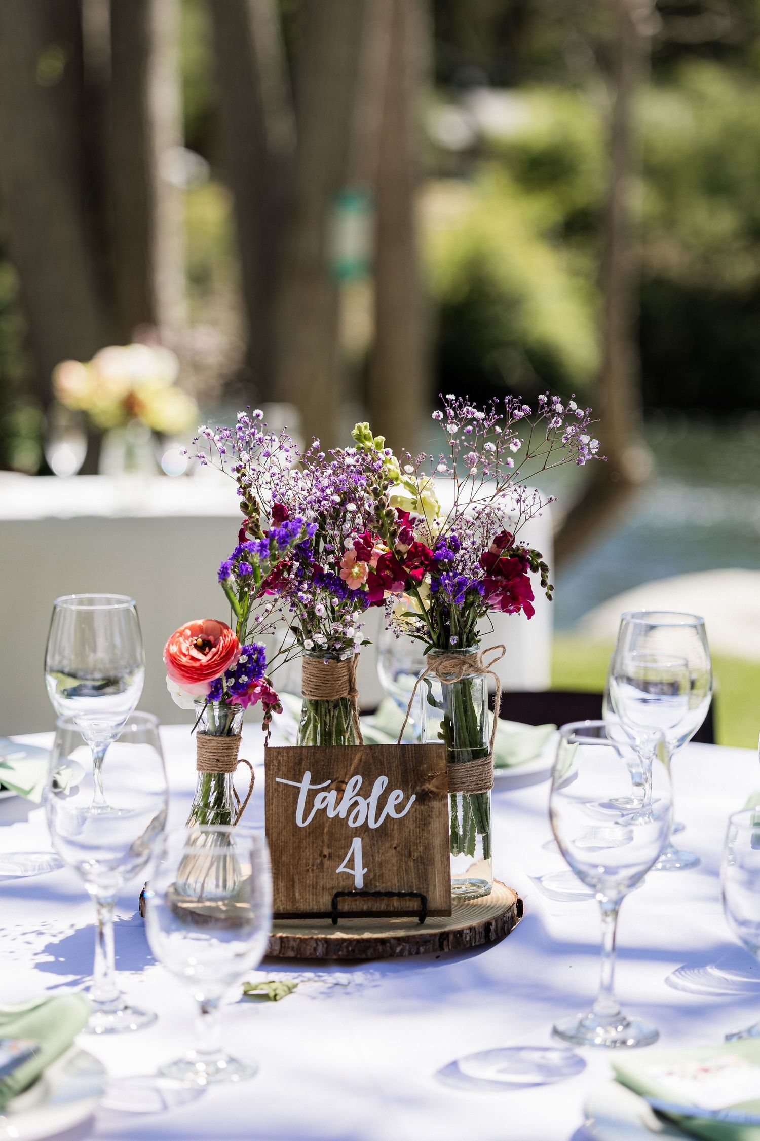 Rustic wedding table setting with wooden box centerpieces filled with colorful wildflowers and wine glasses on white linens.