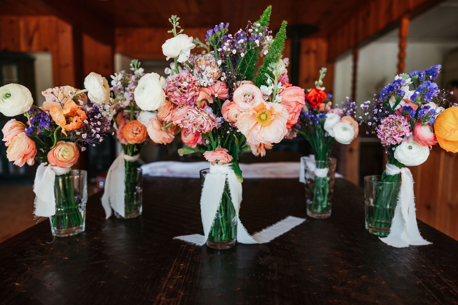 Colorful floral arrangements in glass vases line a rustic wooden table at a wedding reception.
