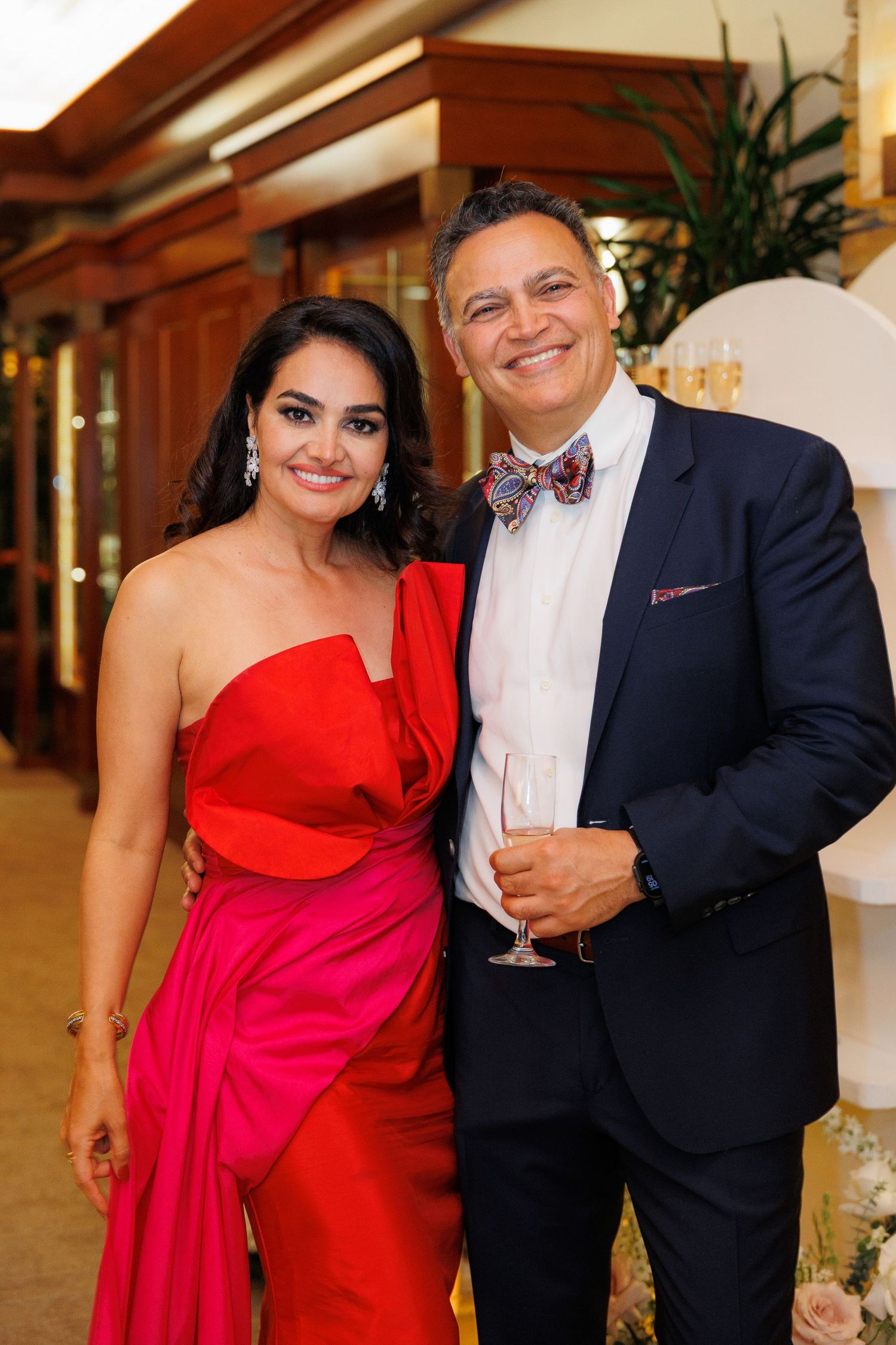 A couple in formal attire poses at an event, with the woman wearing a vibrant red gown and the man in a dark suit.