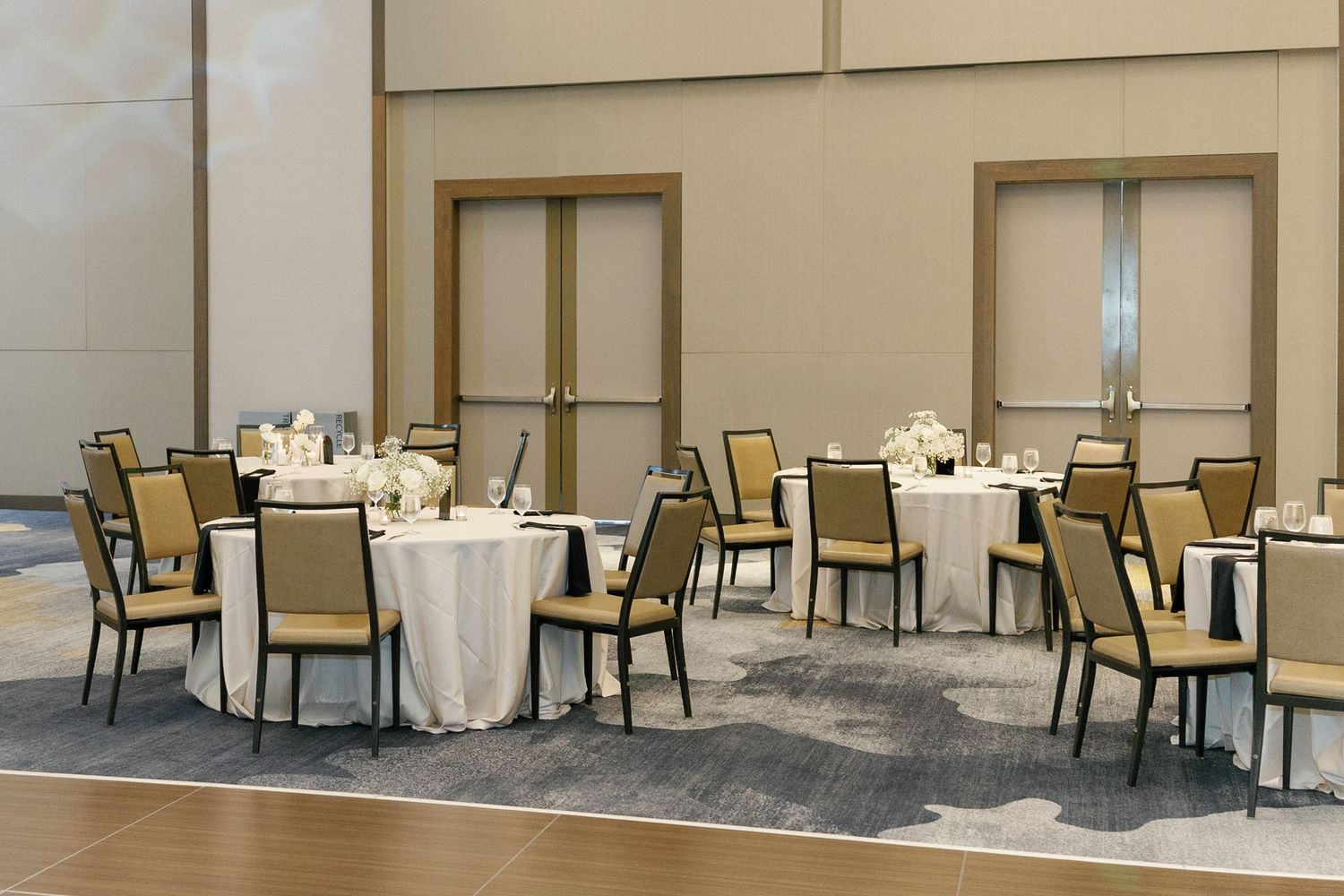 Modern banquet hall setup with round tables, cream tablecloths, and dark wood chairs on patterned carpet.