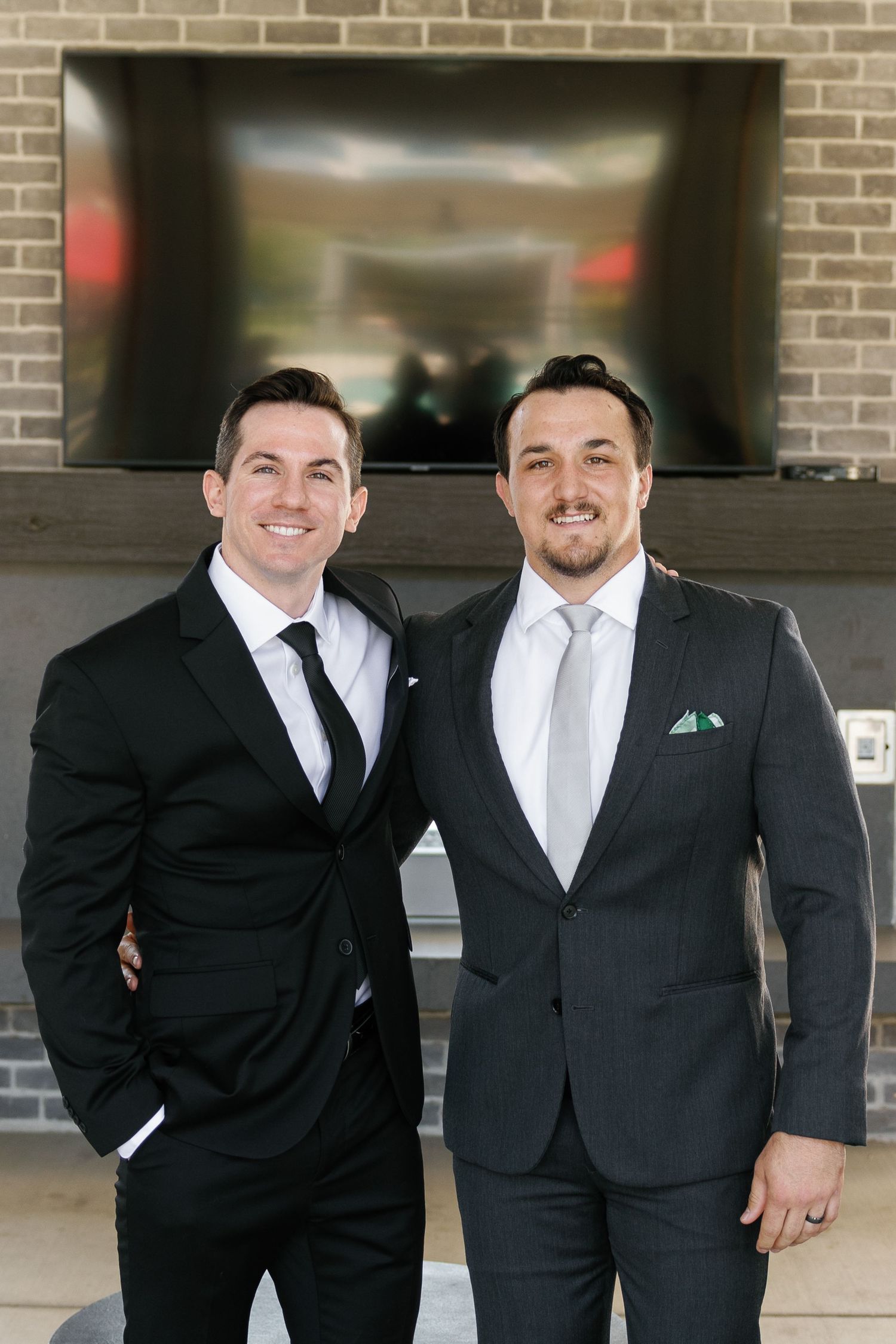 Two people wearing formal black suits and ties pose together against a brick wall with TVs mounted in the background.
