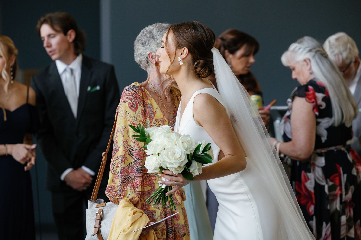 Wedding guests gather at a reception venue as a bride in white dress holds a bouquet of white flowers.