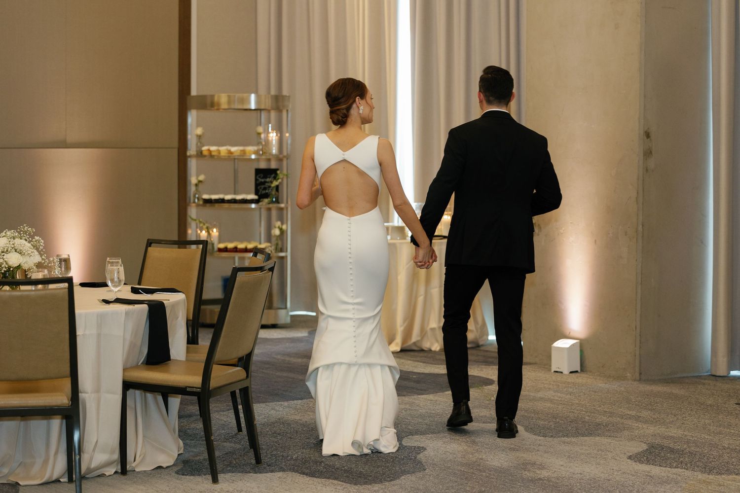 A couple in wedding attire walks away from the camera down an elegant hallway with white walls and ornate decor.