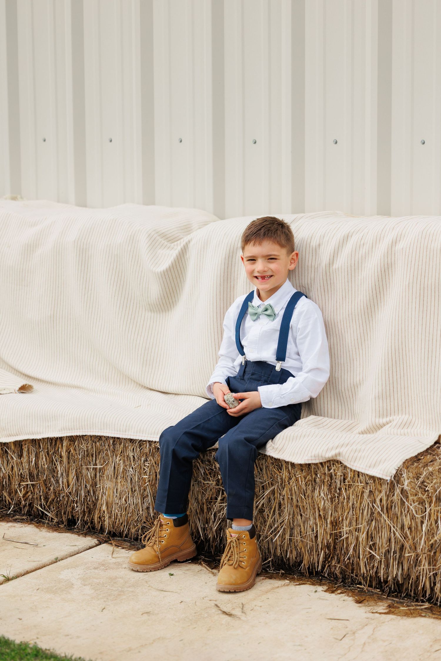A young child in navy overalls and white shirt sits on hay bales against a white backdrop in a rustic photo series.