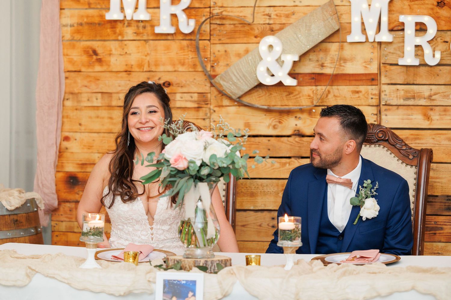 A couple sits at their wedding reception table with rustic wooden decor and 'Mr & Mr' signage on the wall behind them.