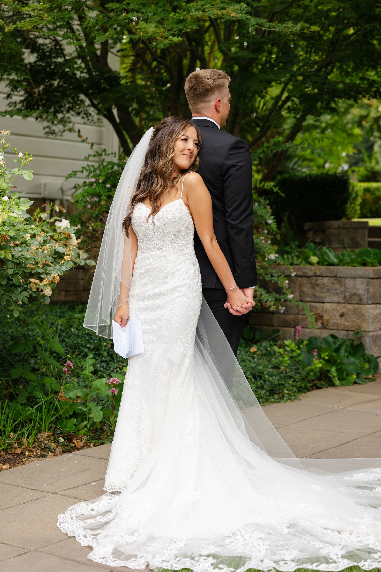 A bride in a white lace wedding gown with a flowing cathedral veil stands on stone steps surrounded by greenery.