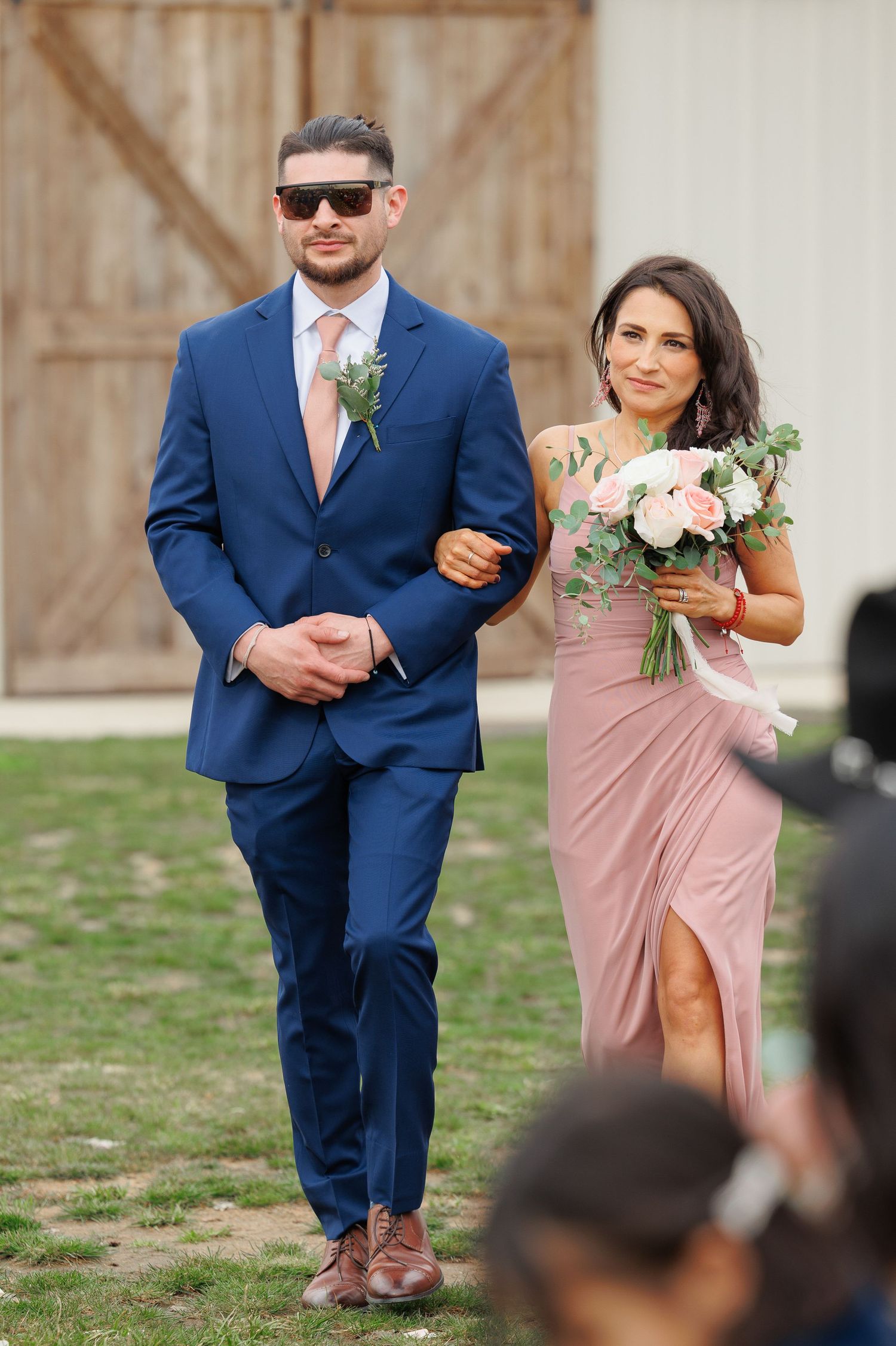 Wedding party members in navy blue suits and blush pink dresses walk down an outdoor aisle by a rustic barn.