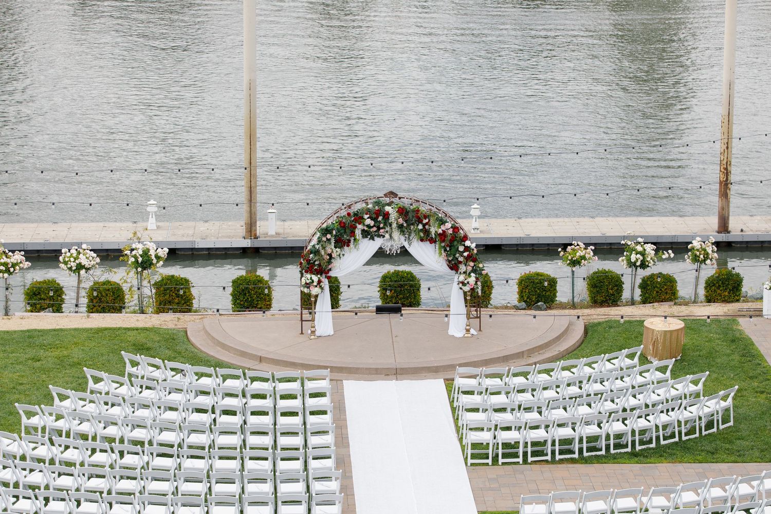 Outdoor wedding ceremony setup with white chairs arranged in curved rows facing a floral arch by the water.