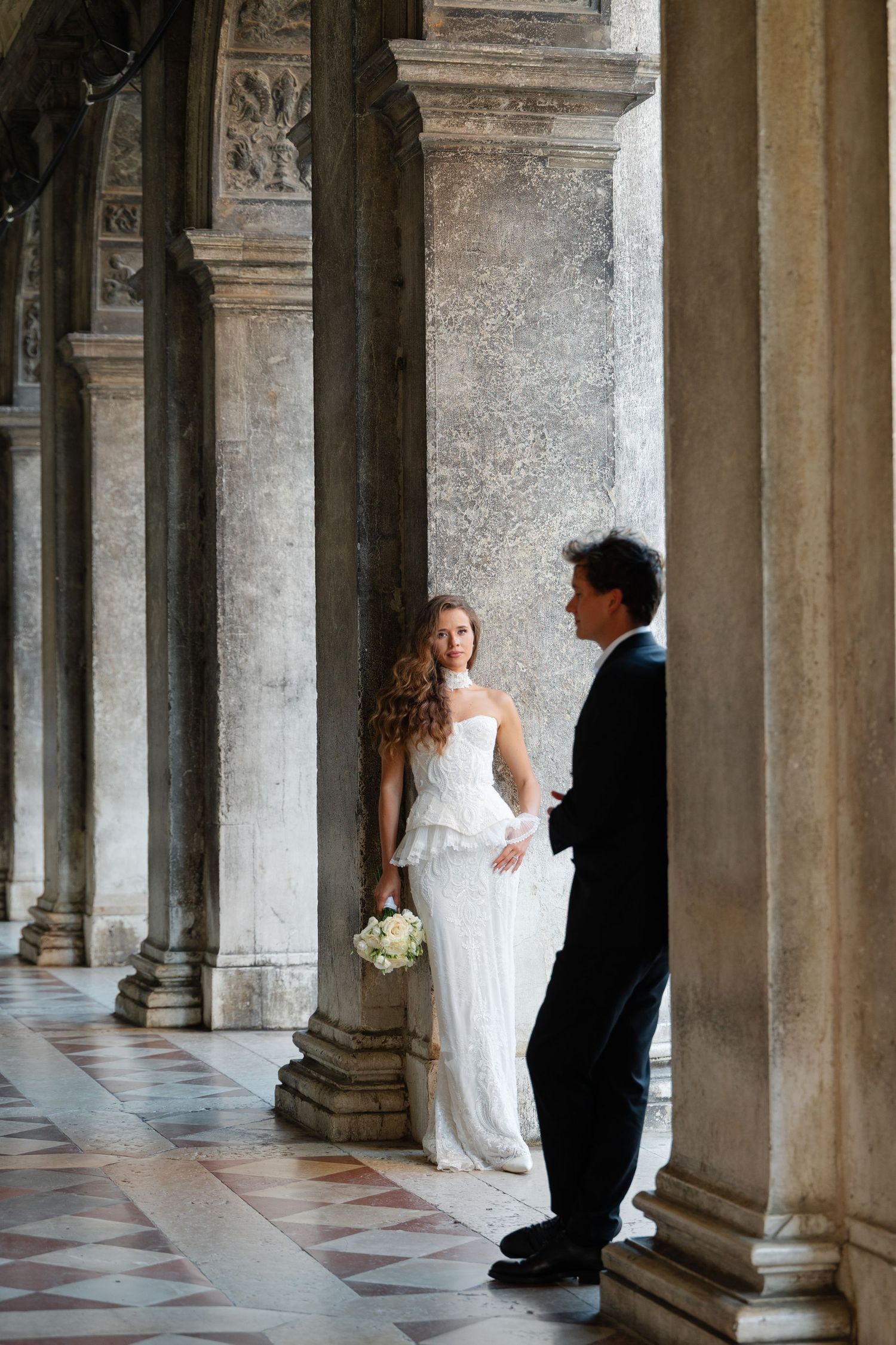 Elegant wedding couple poses between ornate stone columns in a historic architectural colonnade.