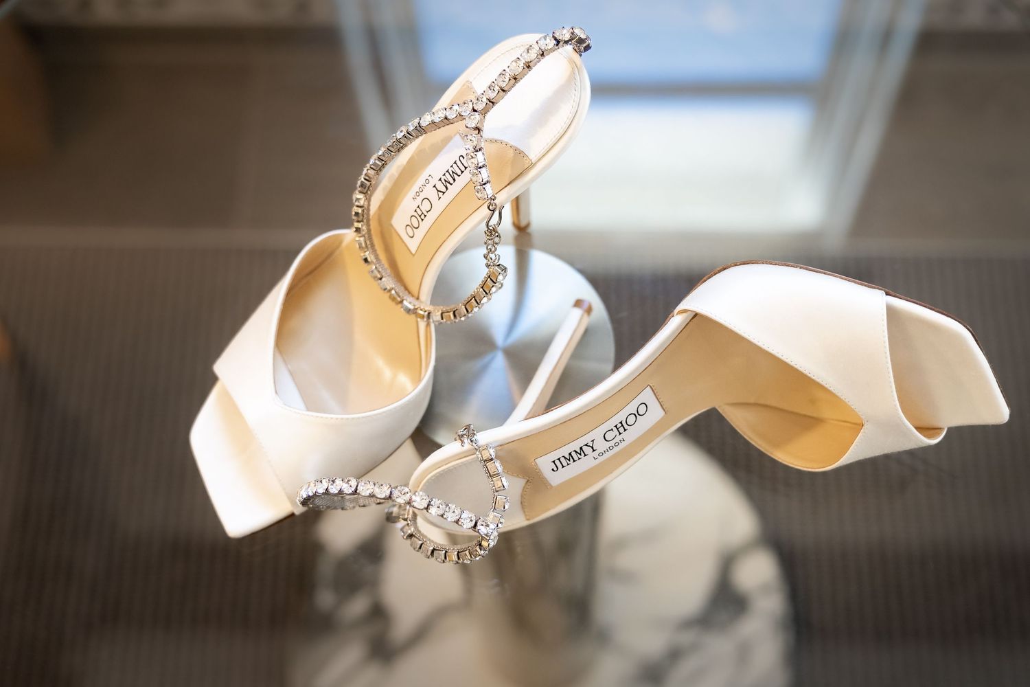 White satin wedding shoes with crystal embellishments and delicate bow details sit elegantly on a reflective surface.