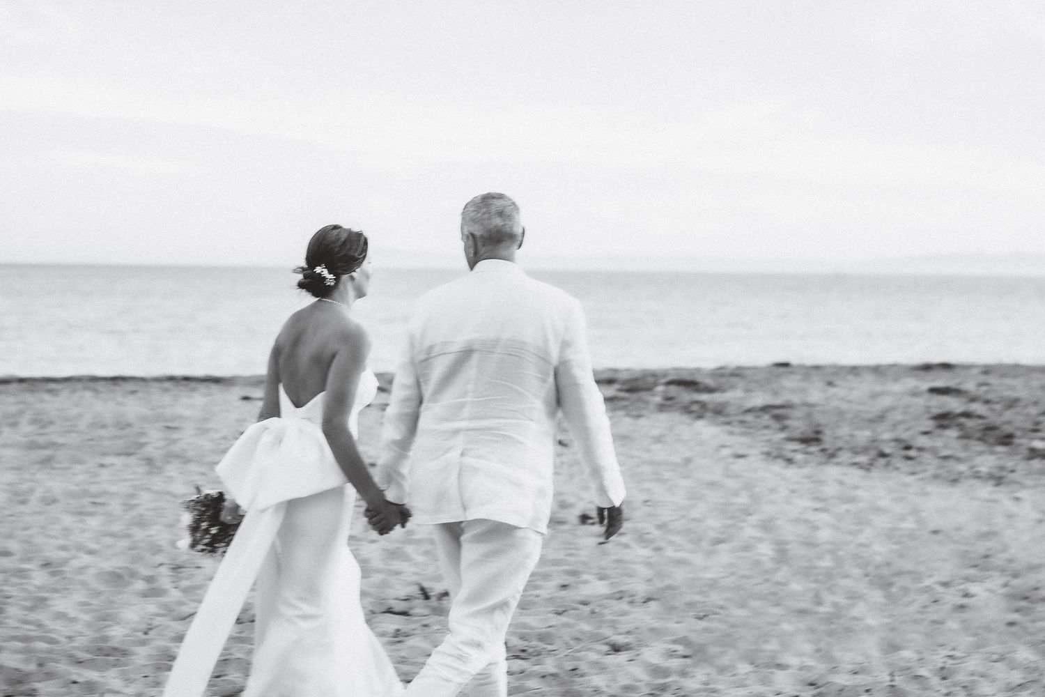 A couple walks hand in hand along a beach shore in black and white.