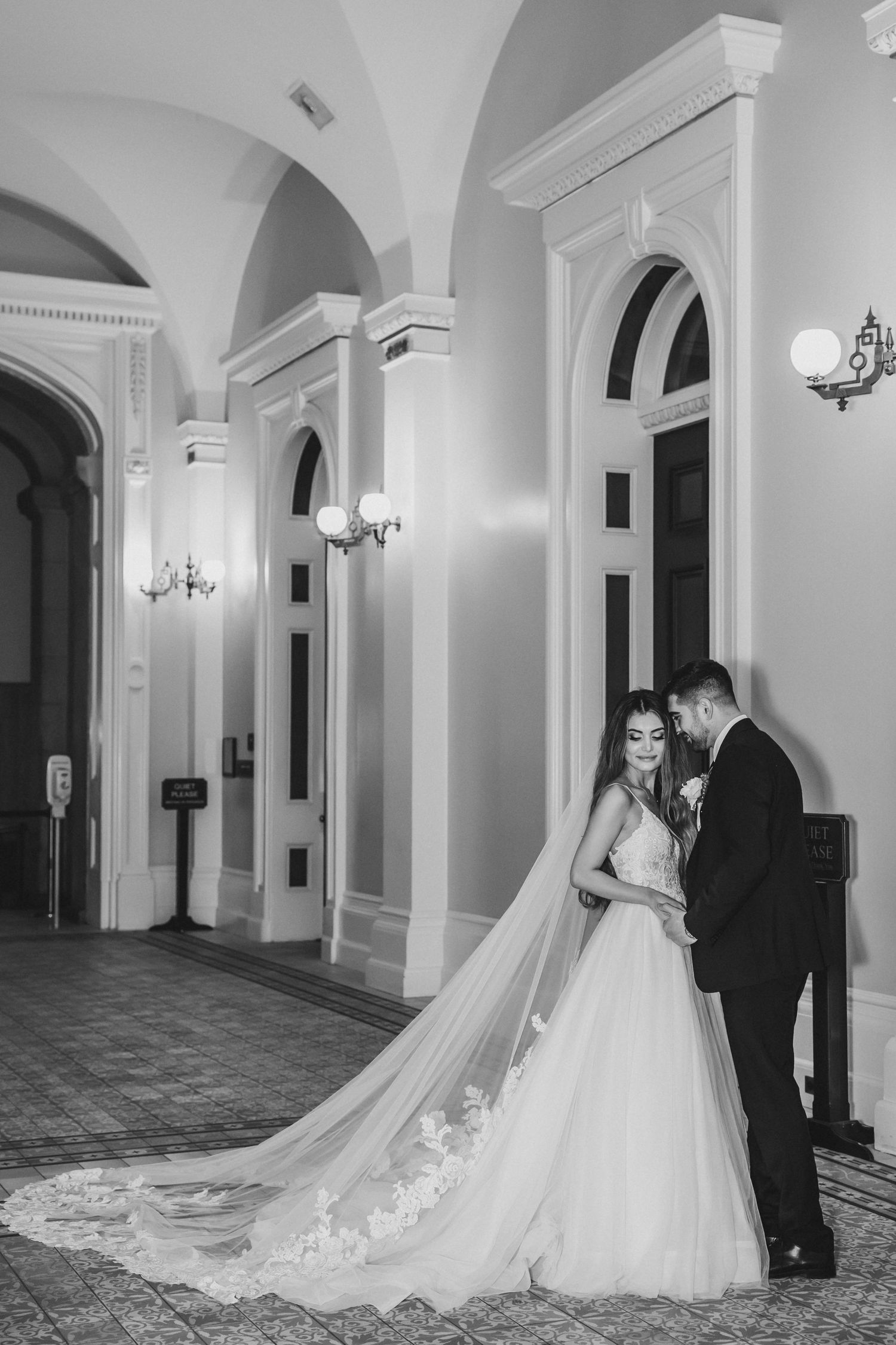 Black and white wedding photo of bride and groom in elegant hall with arched ceilings and long lace veil.
