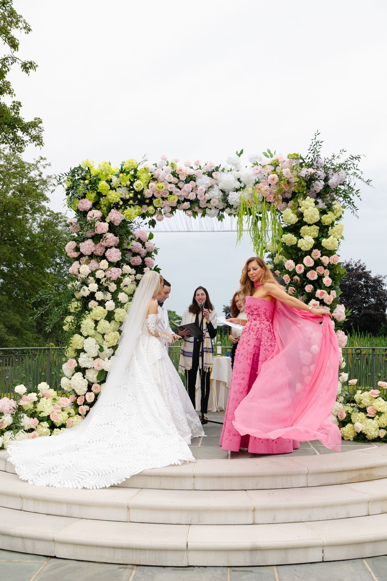 A wedding party poses on stone steps under a floral arch decorated with white and yellow roses at an outdoor ceremony.