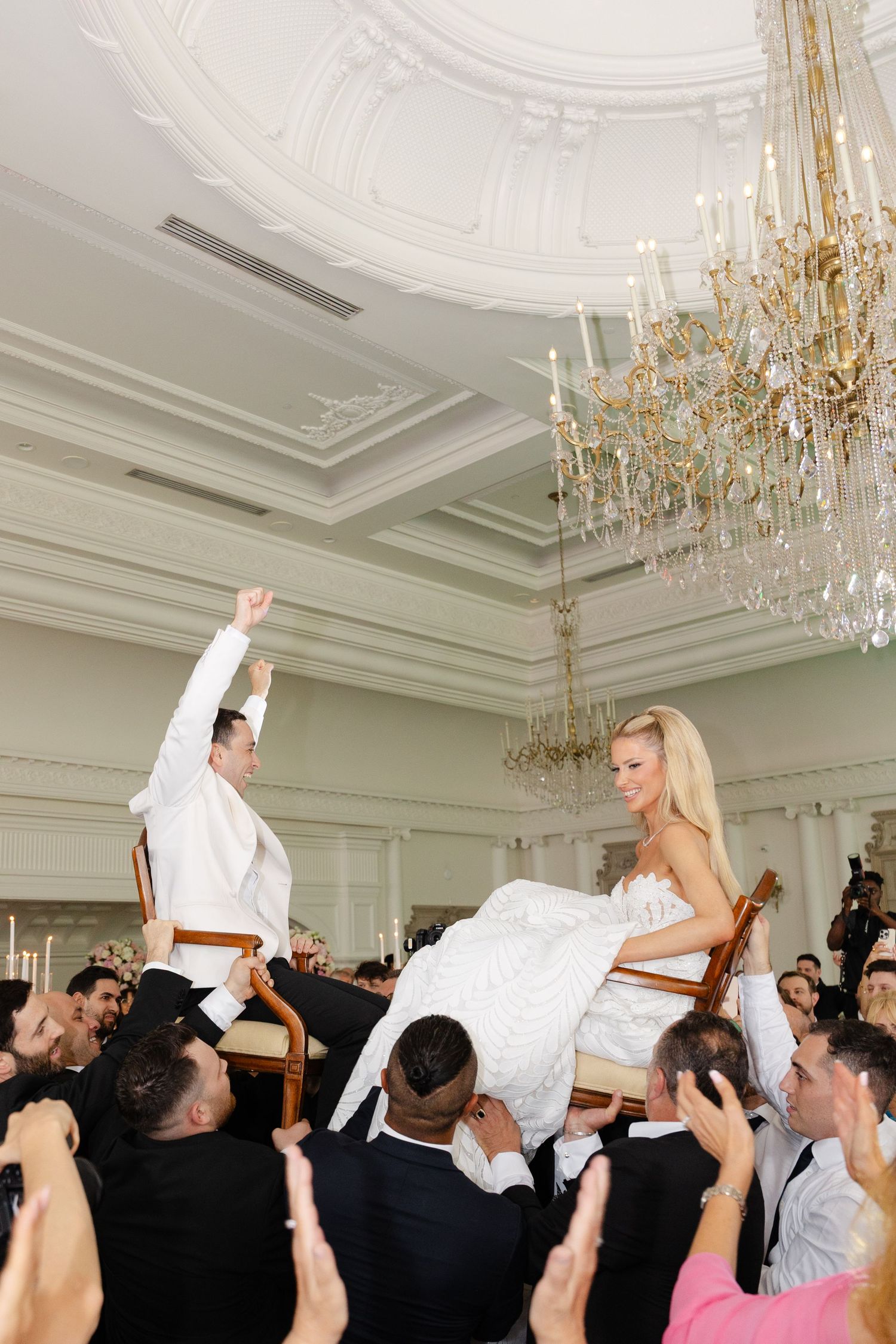 Wedding guests lift and toss newlyweds during a reception celebration in an elegant ballroom with crystal chandeliers.