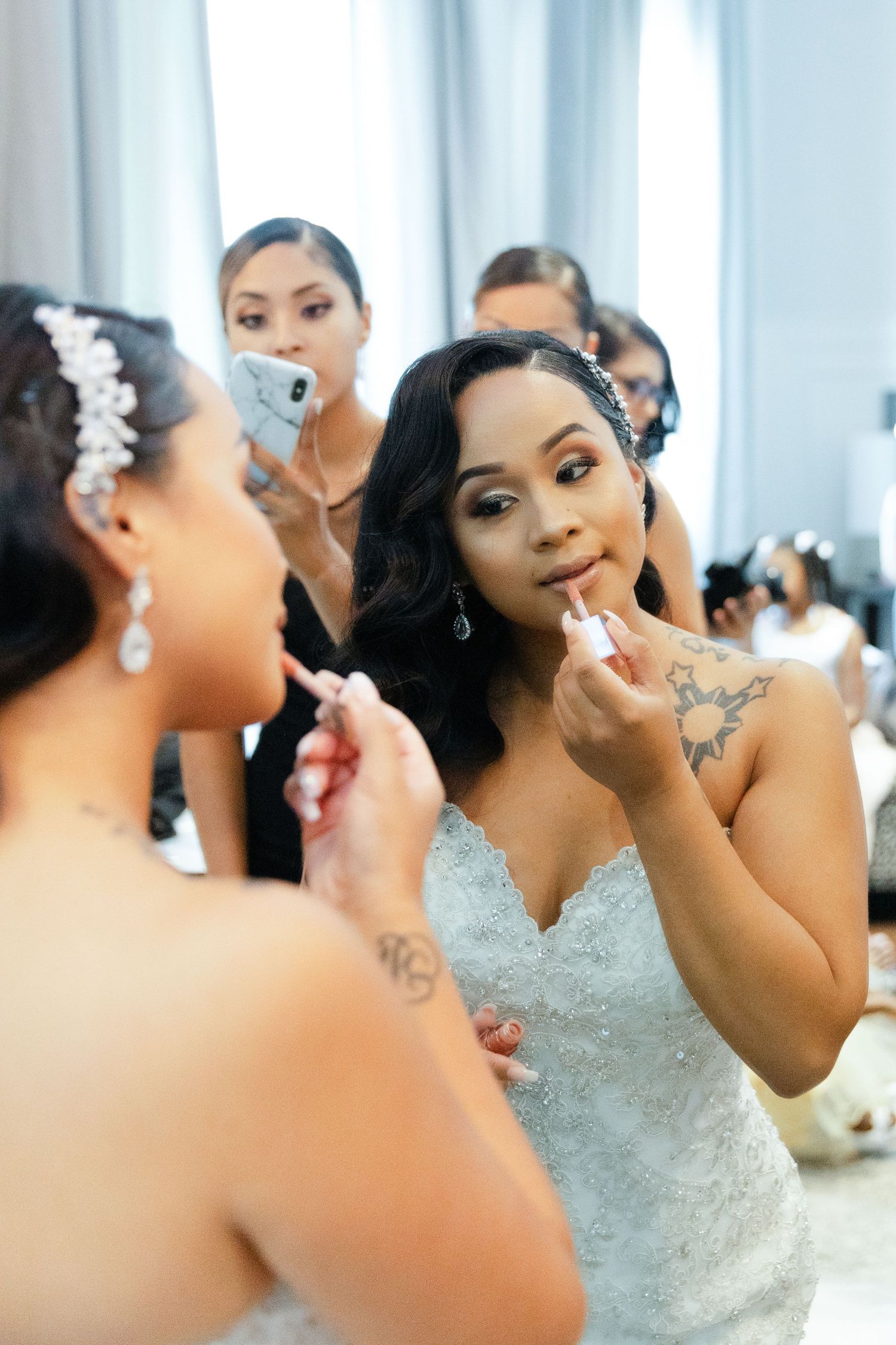 A bride in a white lace gown applies lipstick while getting ready with her bridal party in a bright room.