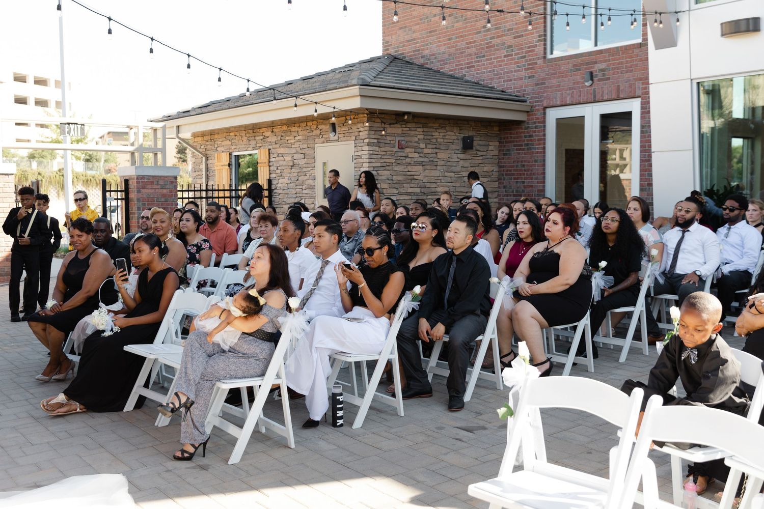 Panoramic view of wedding guests seated in white chairs at an outdoor ceremony venue with brick walls.