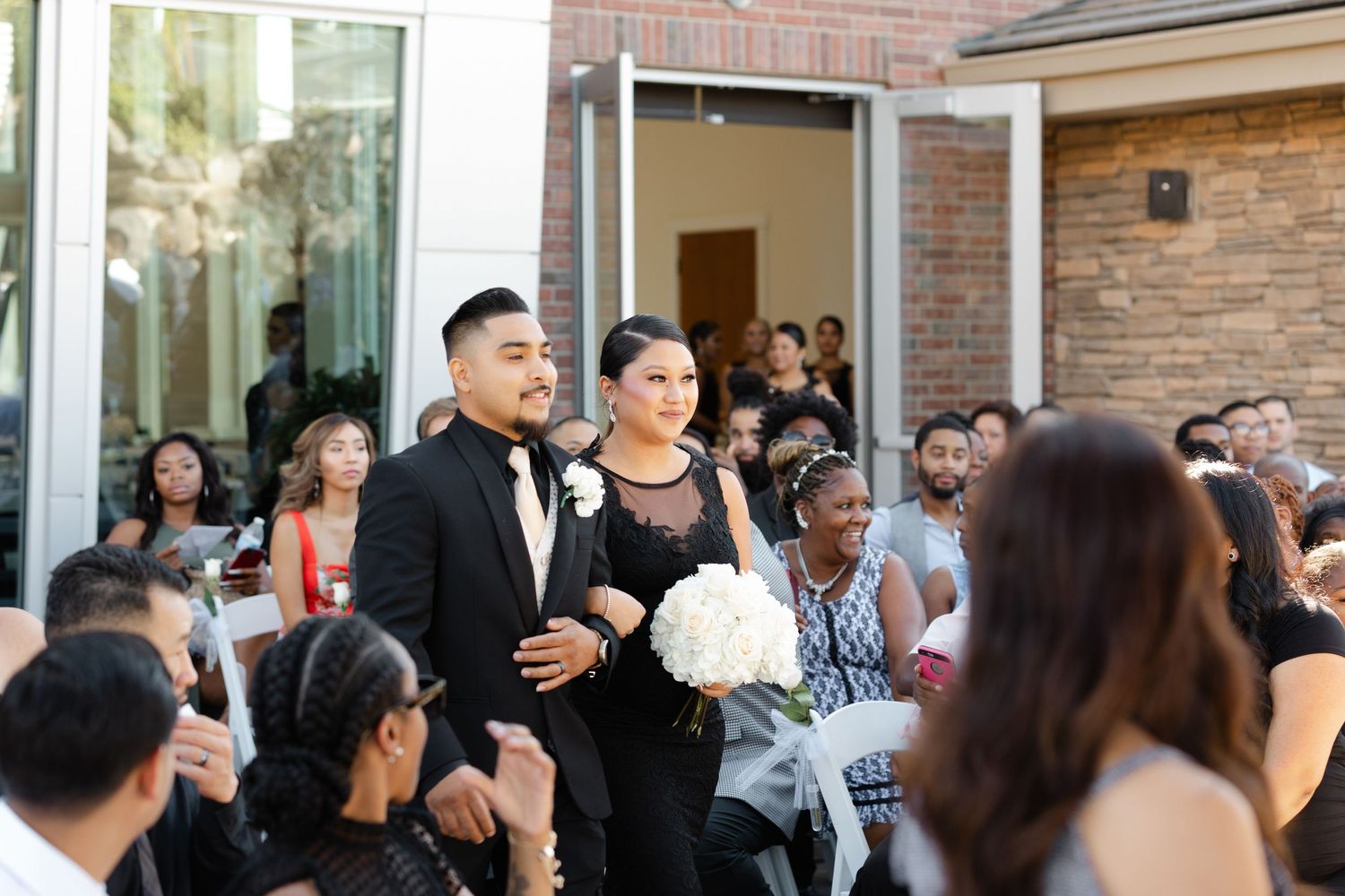 Wedding party members walk down the aisle during an outdoor ceremony while guests look on at a brick venue.