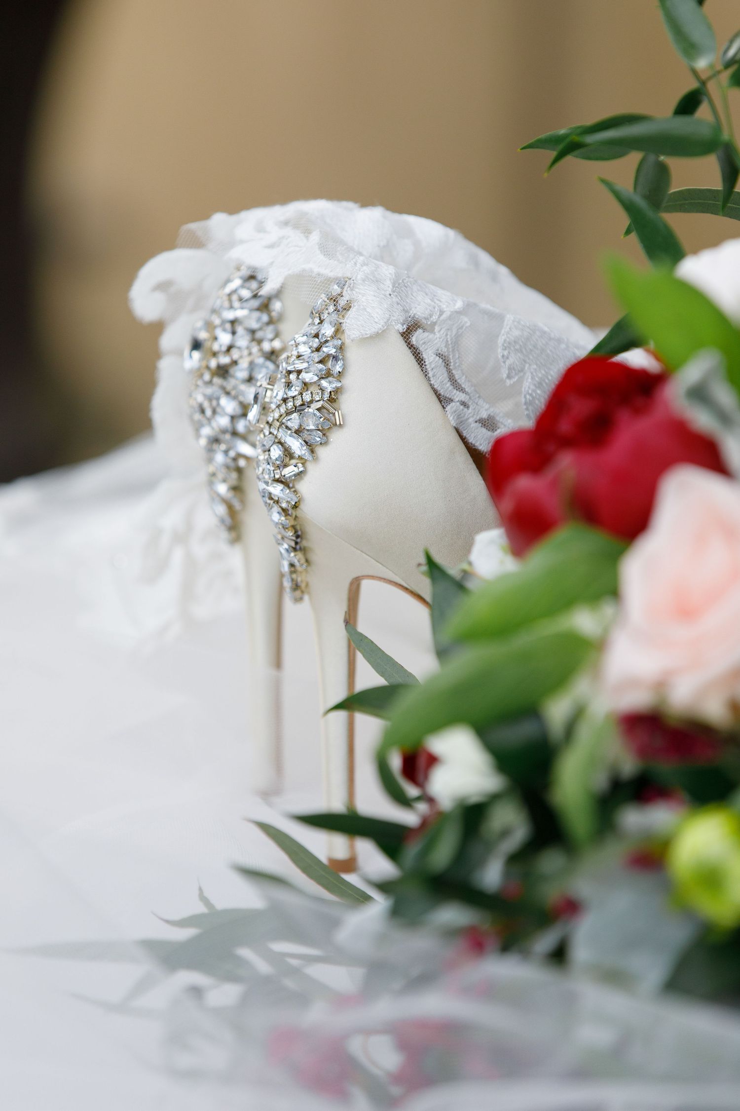 Close-up detail of a white wedding shoe with sparkling crystal embellishments surrounded by soft floral decorations.