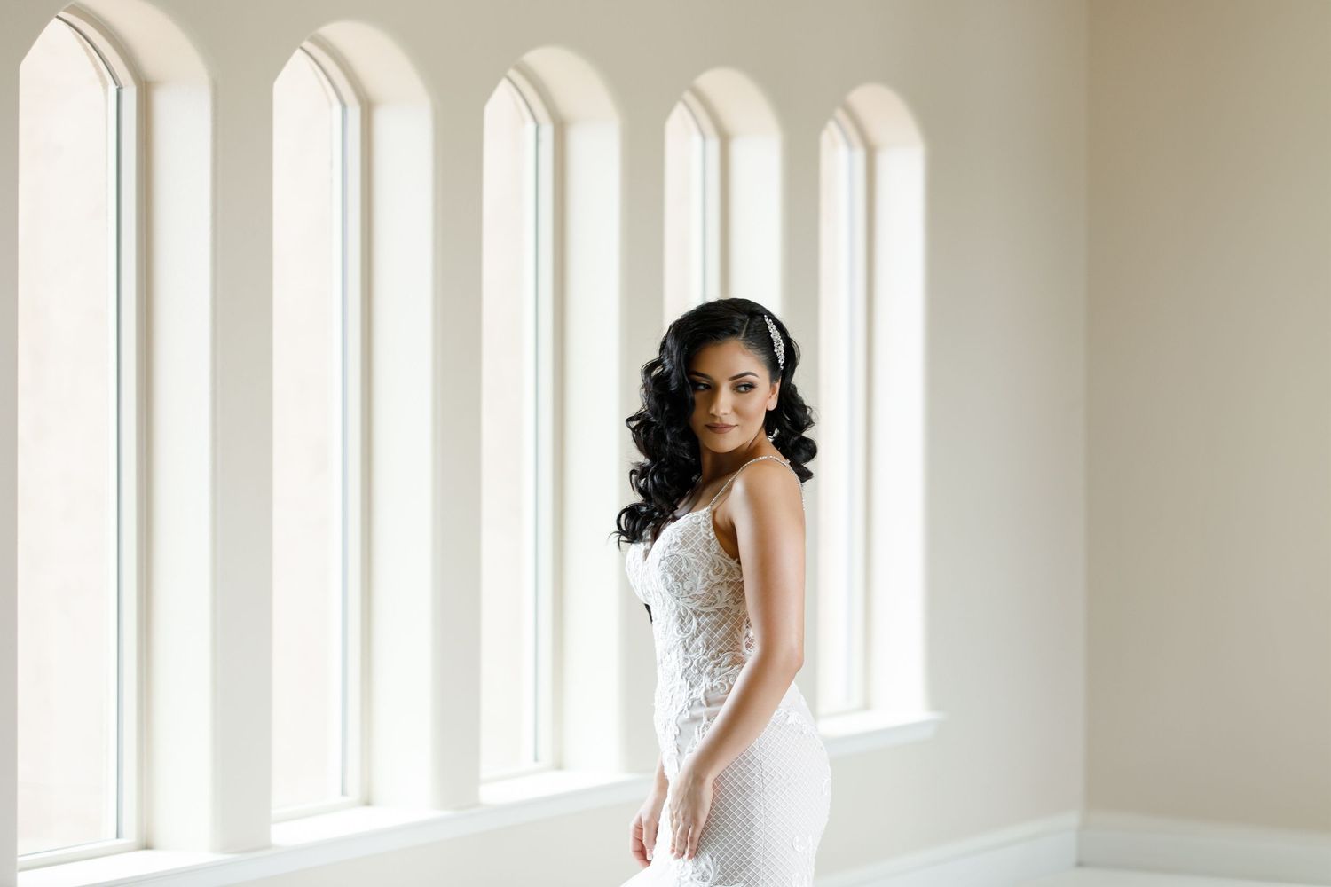 A person in a white dress poses gracefully by a row of bright windows in an elegant interior setting.