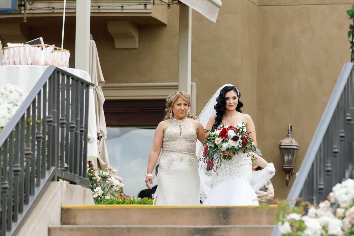Two people in elegant wedding attire pose on a grand staircase with floral arrangements and classic architecture.