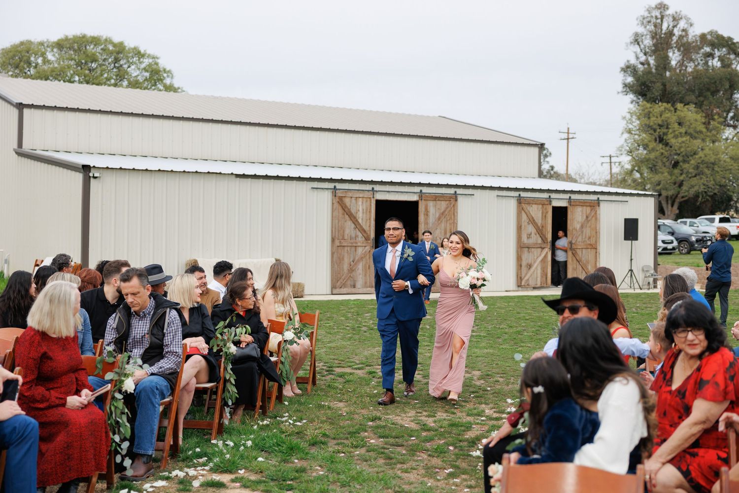 Wedding guests gather outdoors on a grassy area near white buildings during a sunny day celebration.
