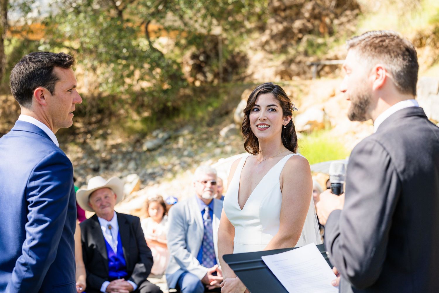 An outdoor wedding ceremony takes place in a sunlit natural setting with guests gathered to witness the occasion.