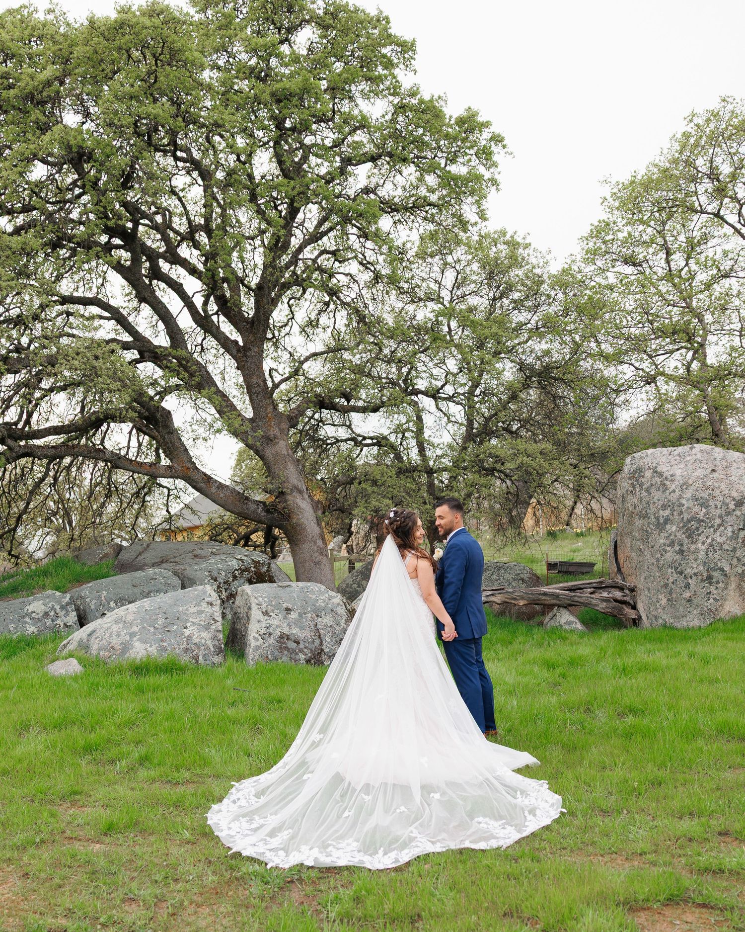 A couple in wedding attire poses in a scenic outdoor setting with large oak trees and boulders in the background.