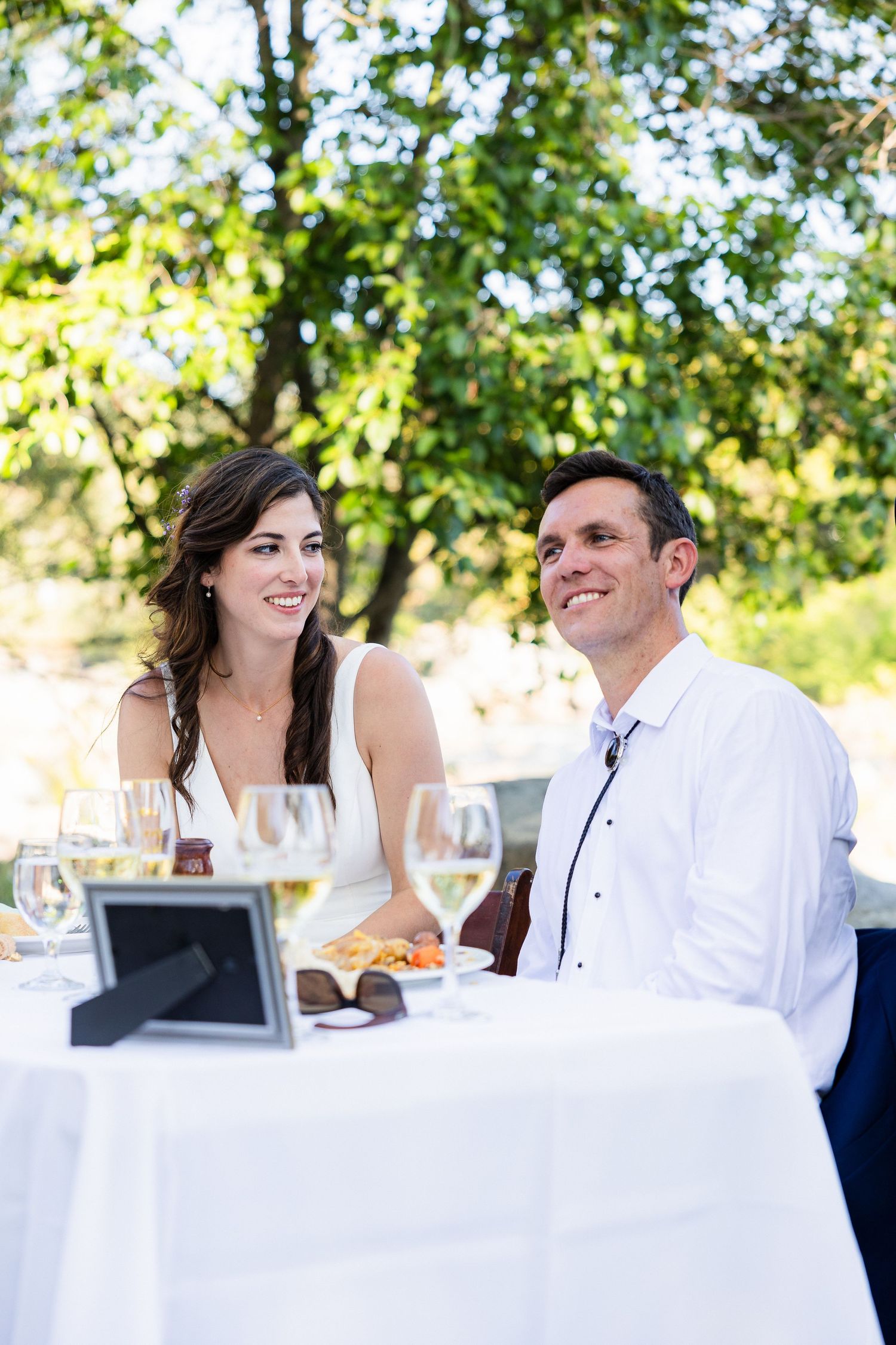 A couple enjoys an elegant outdoor dining experience at a white-clothed table with wine glasses in a garden setting.