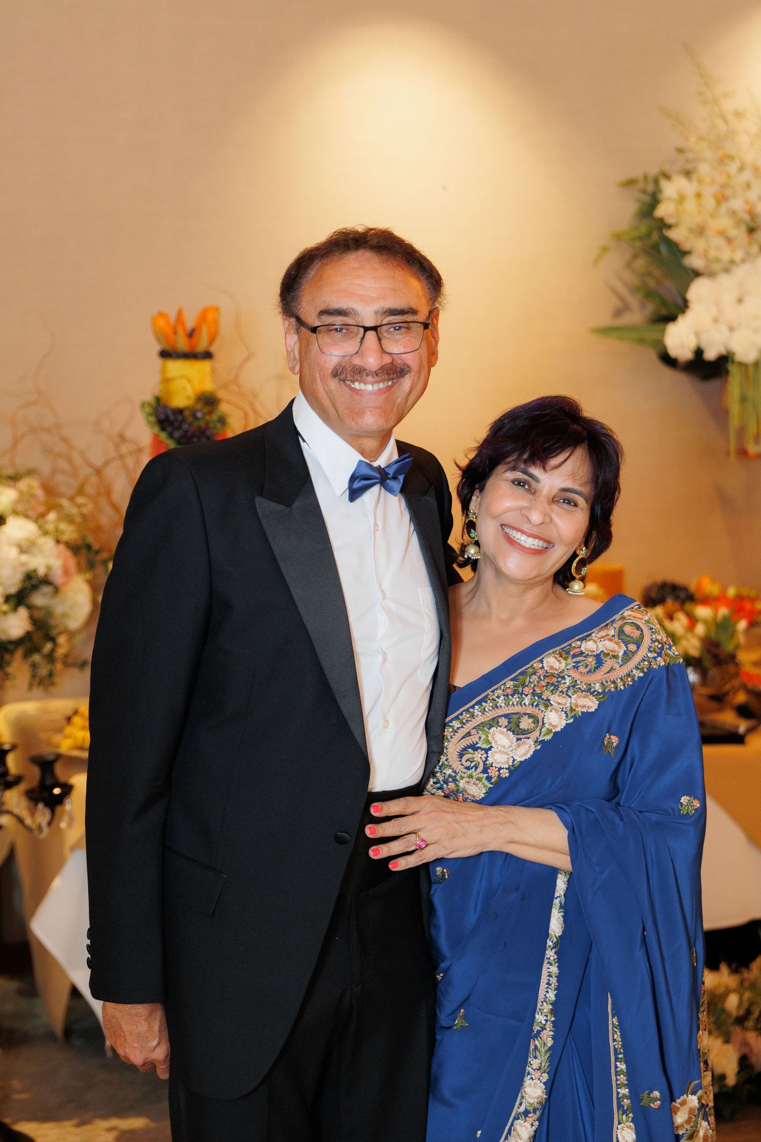 A couple poses together at a formal event, with the woman wearing an elegant blue and gold embroidered dress.