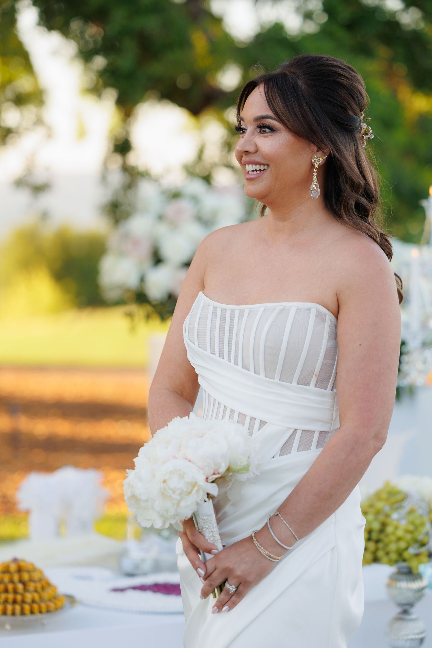 A bride in a white strapless wedding dress smiles while sitting at an outdoor reception table during golden hour.