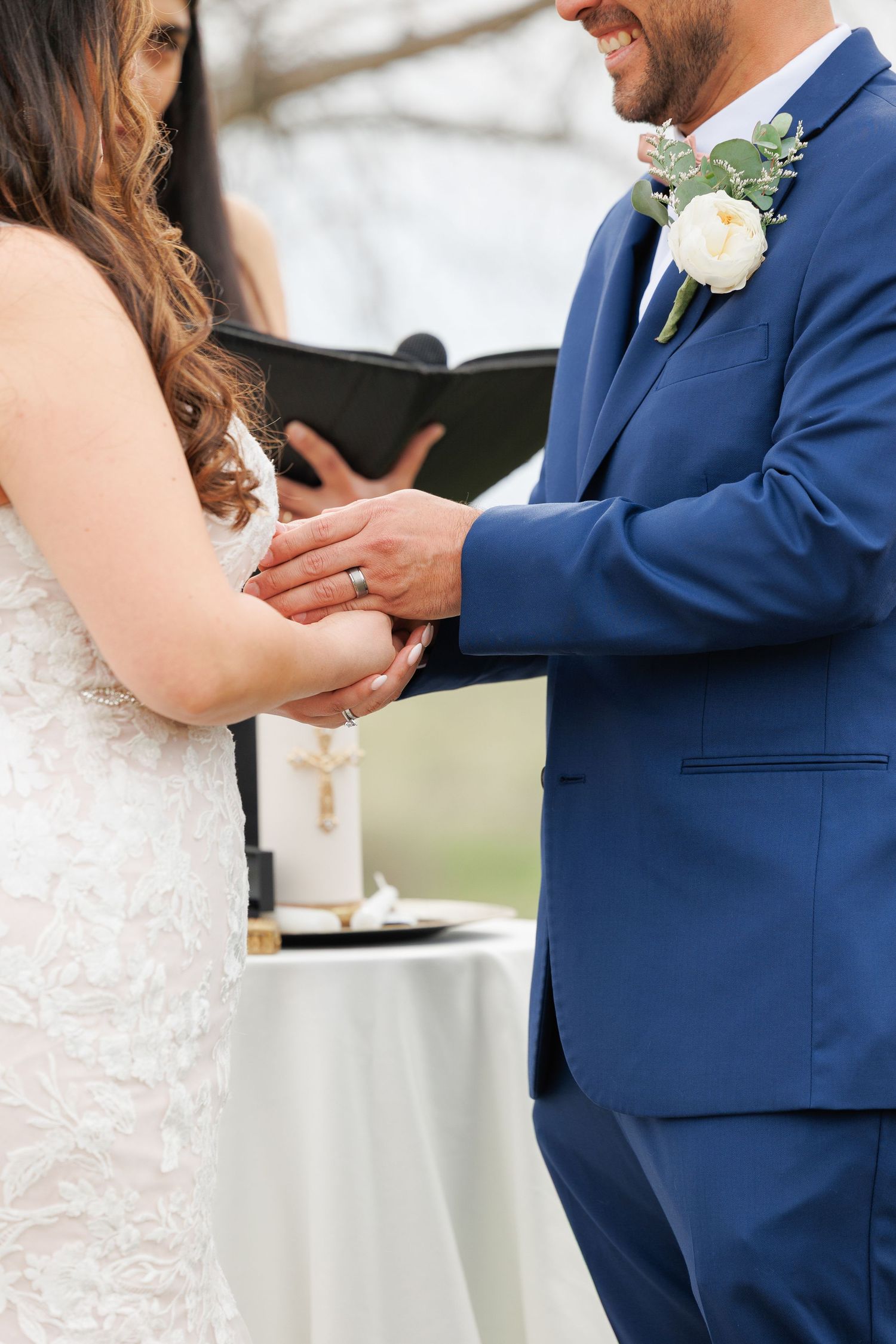 Close-up of bride and groom exchanging rings during outdoor wedding ceremony with white and blue attire.