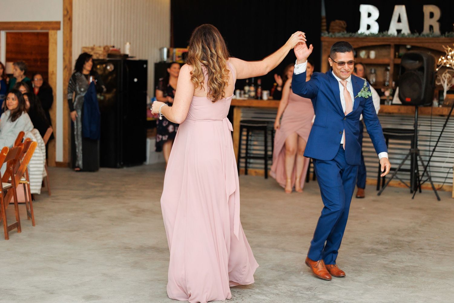 Wedding guests dance and socialize on a rustic barn dance floor with a 'BAR' sign visible in the background.