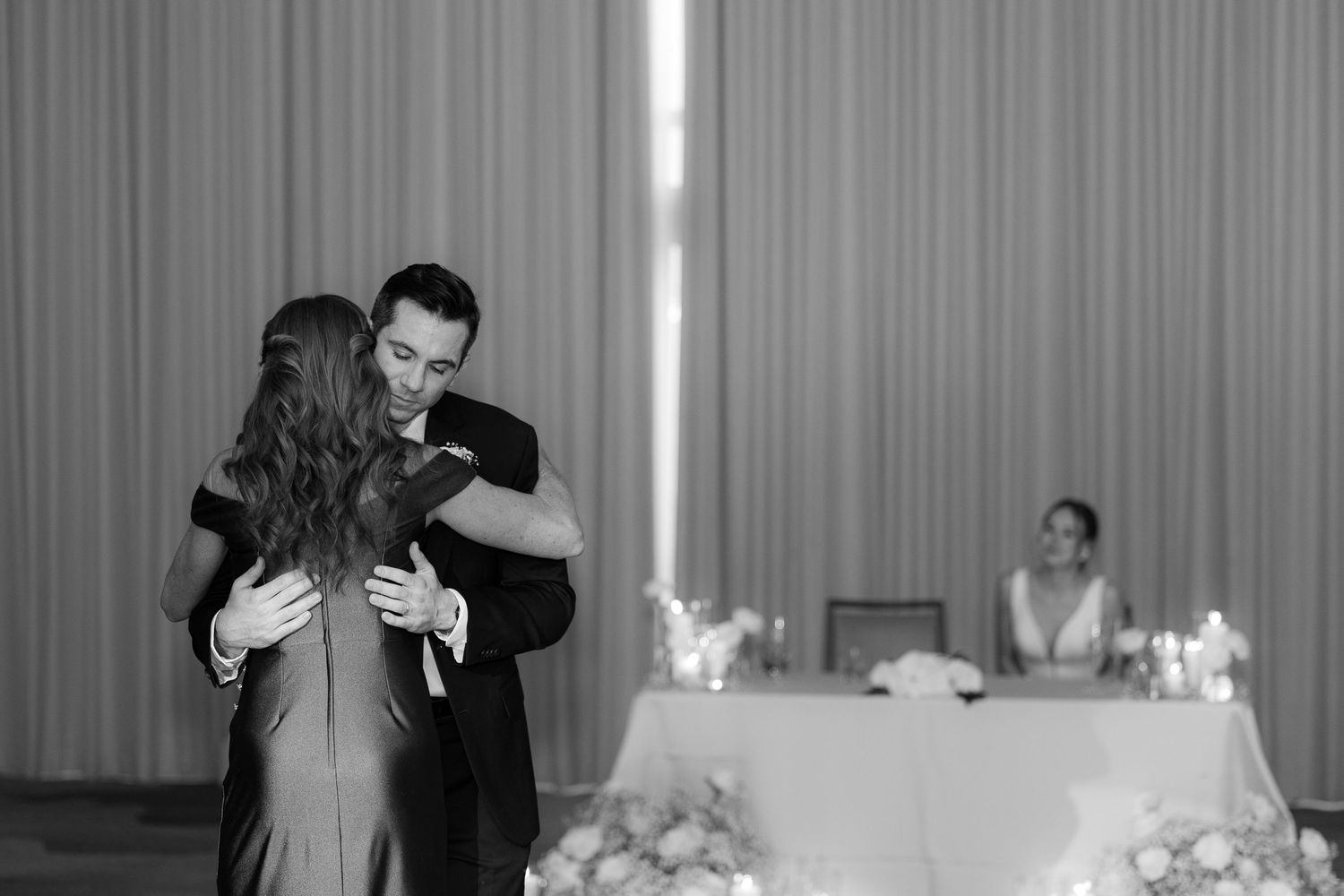 A wedding reception scene with guests dancing and celebrating in a banquet hall with white curtains and decor.