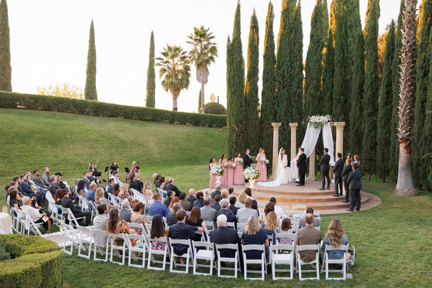 Outdoor wedding ceremony setup with rows of white chairs and tall cypress trees lining the venue.