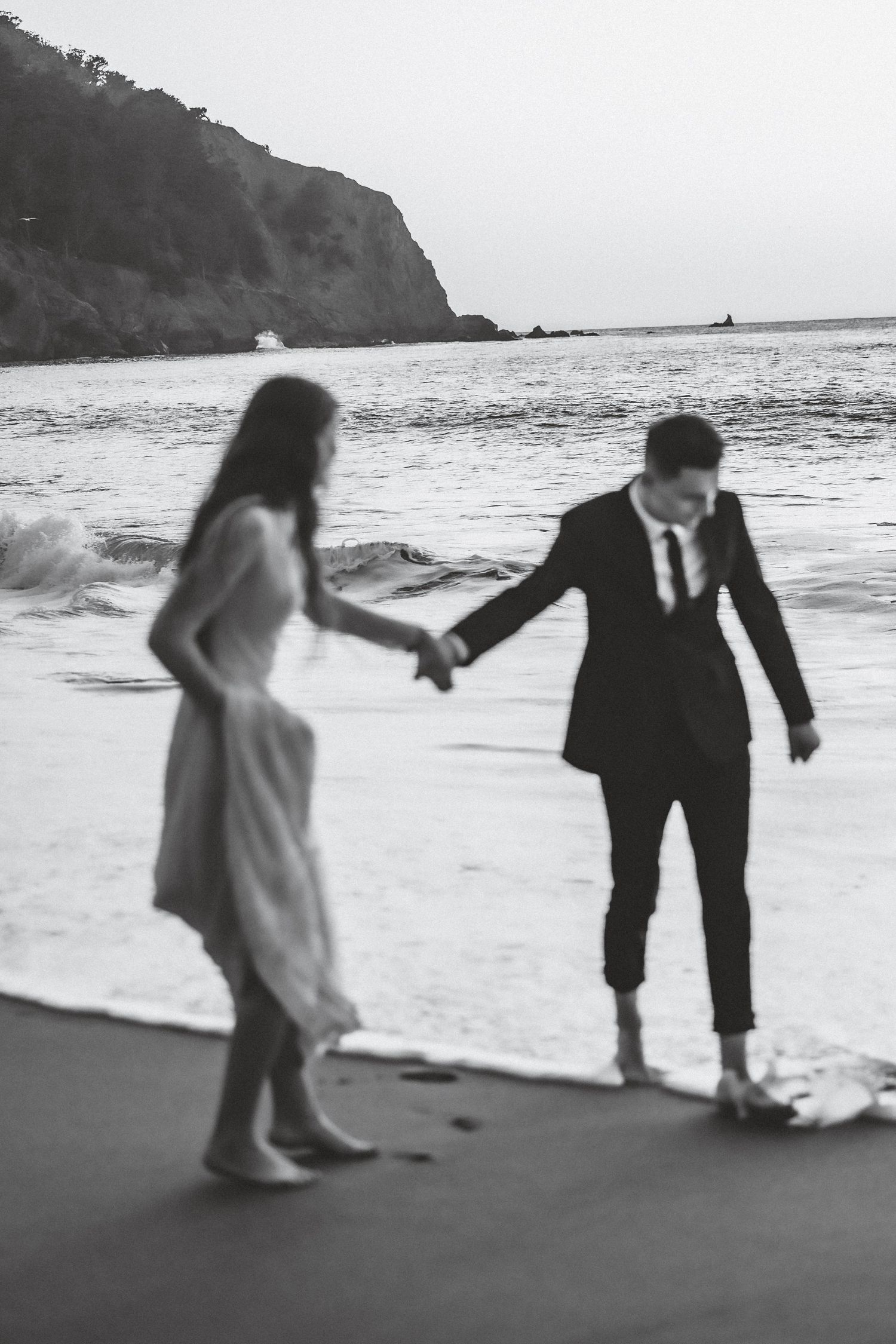 Black and white photo of couple playing and dancing together on a beach with cliffs in background.