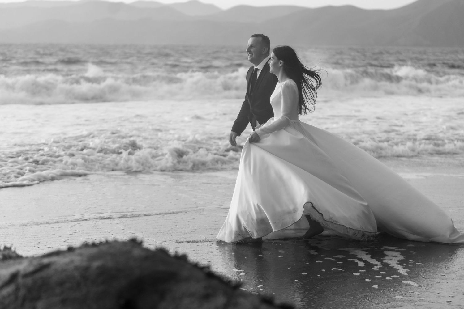 Couple in wedding attire walks along beach shore with waves crashing in black and white.