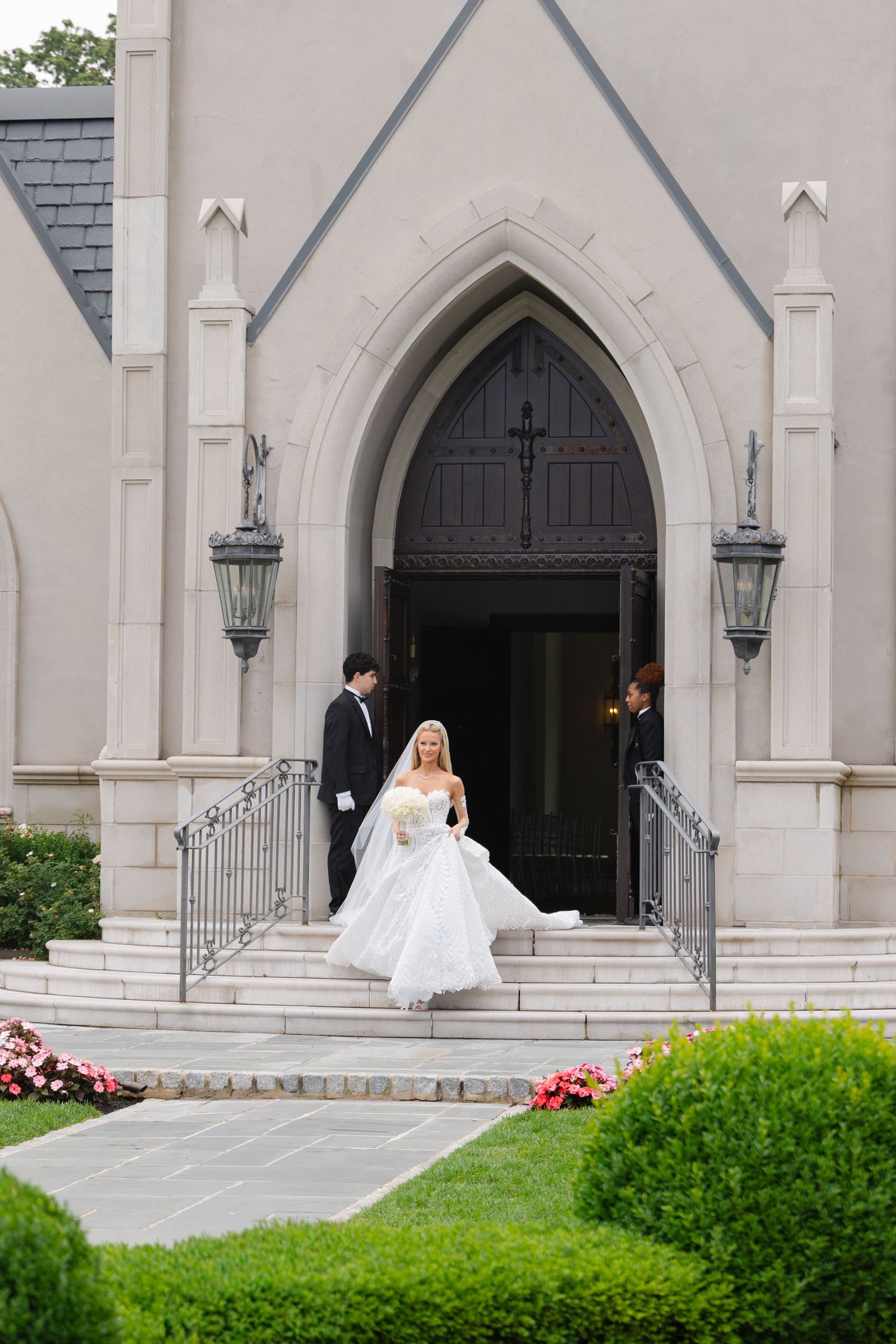 Bride in white gown descends church steps with groom after wedding ceremony at elegant stone chapel.
