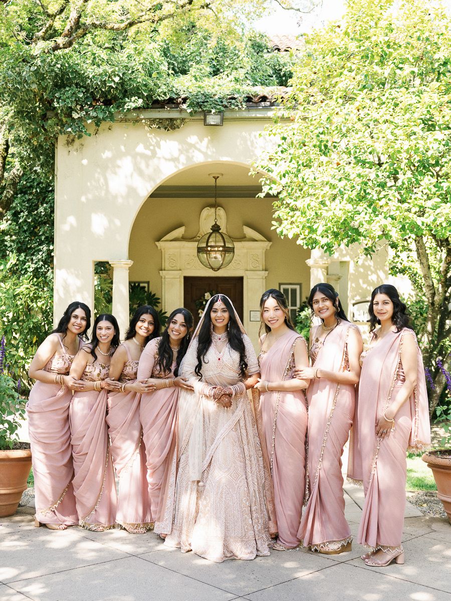 A group wearing matching pink saris poses elegantly in front of an arched entryway covered in lush greenery.
