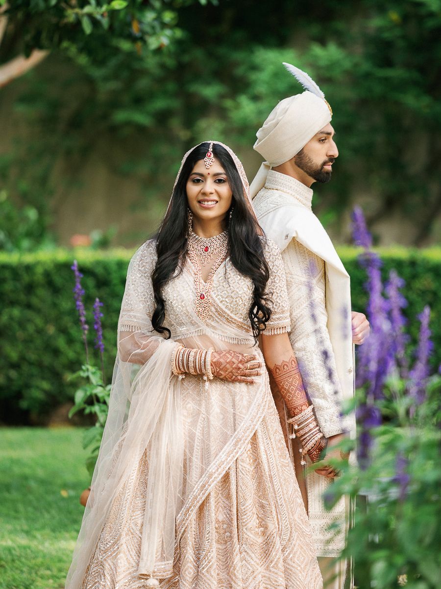 Couple in elegant South Asian wedding attire posing in garden with purple flowers.
