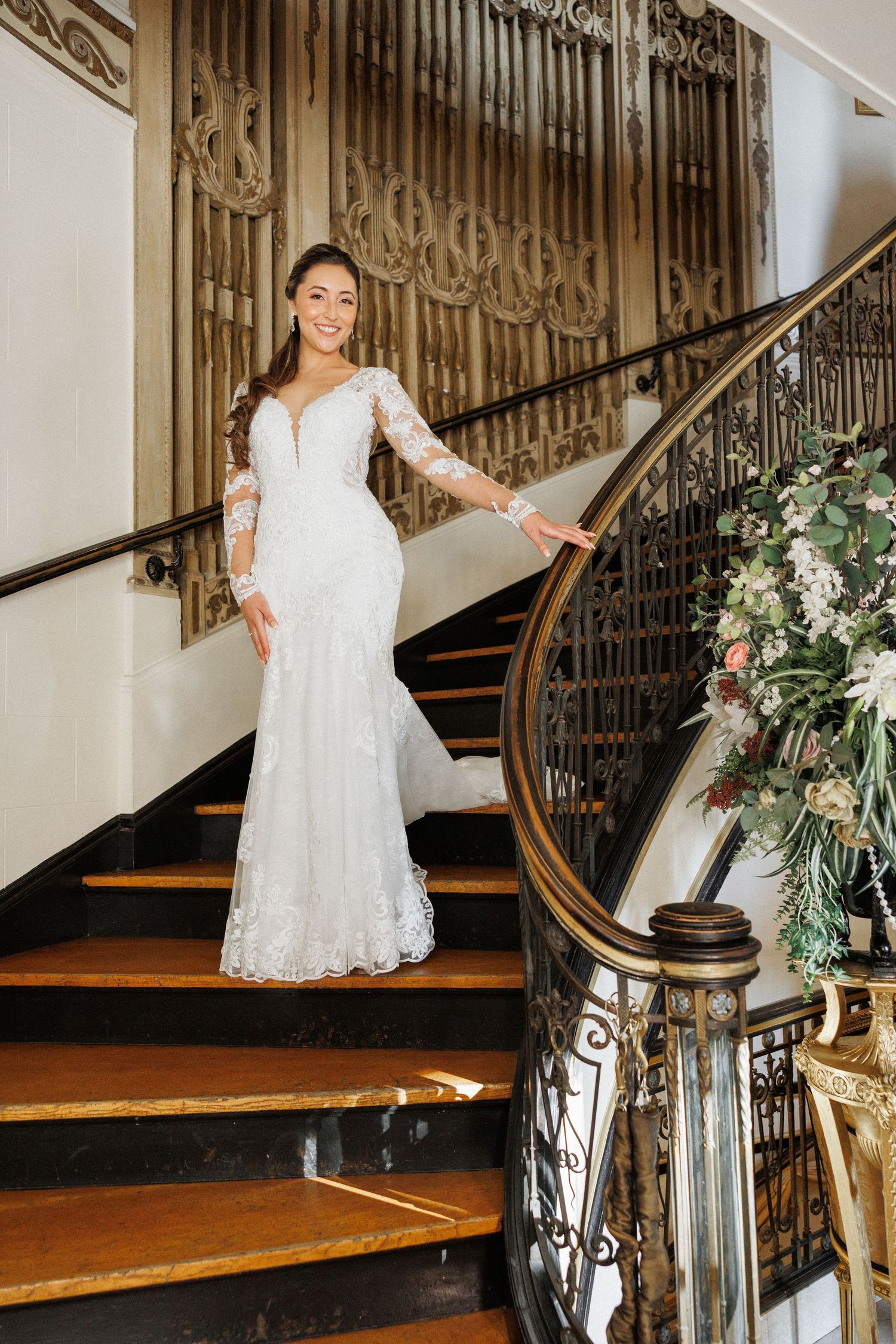 A bride in a lace dress poses on an elegant staircase decorated with floral arrangements.