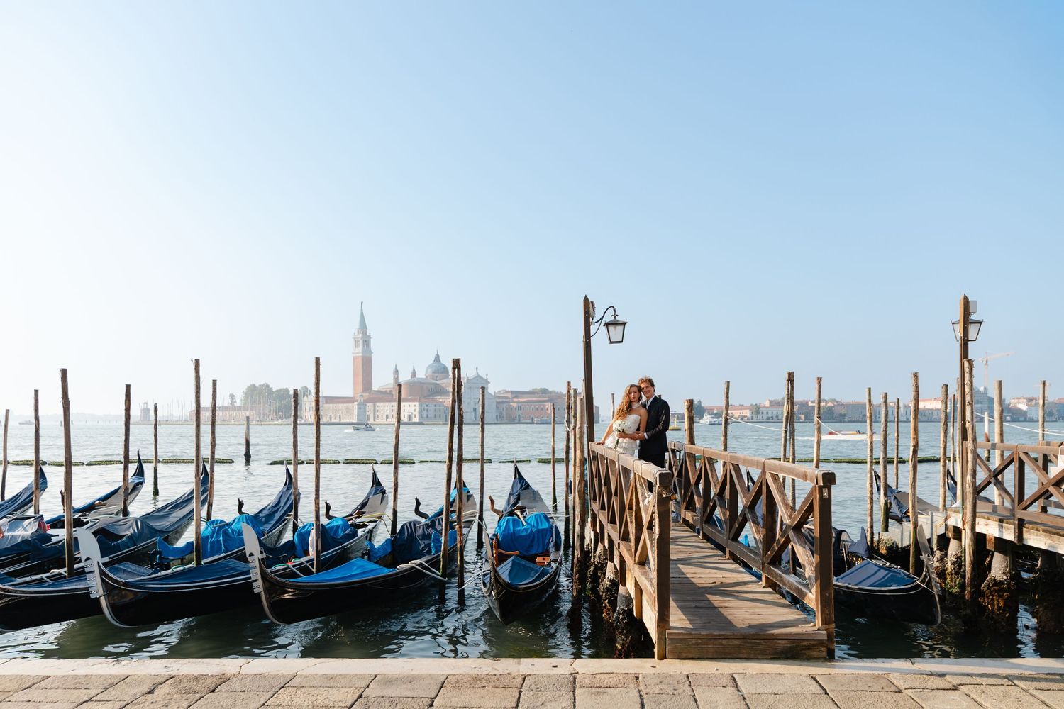 Row of traditional Venetian gondolas moored along wooden posts in Venice lagoon with San Giorgio Maggiore church in background.