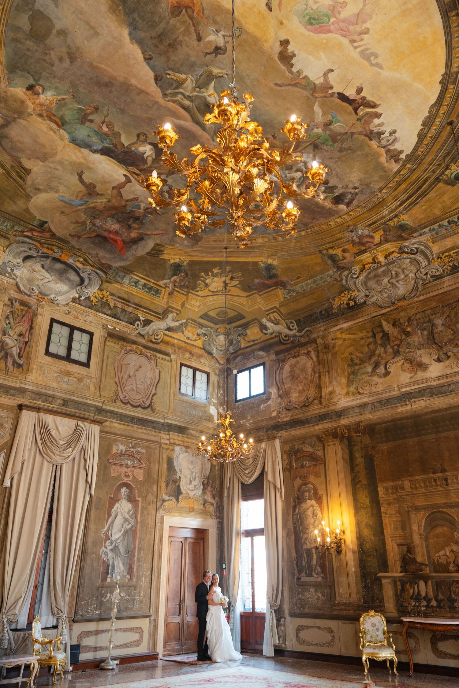 Ornate baroque palace room with gilded chandeliers, frescoed ceilings, and classical statues adorning the grand interior.