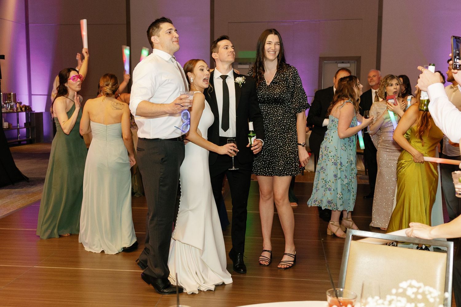 Wedding guests dance together on a purple-lit dance floor during an evening reception celebration.