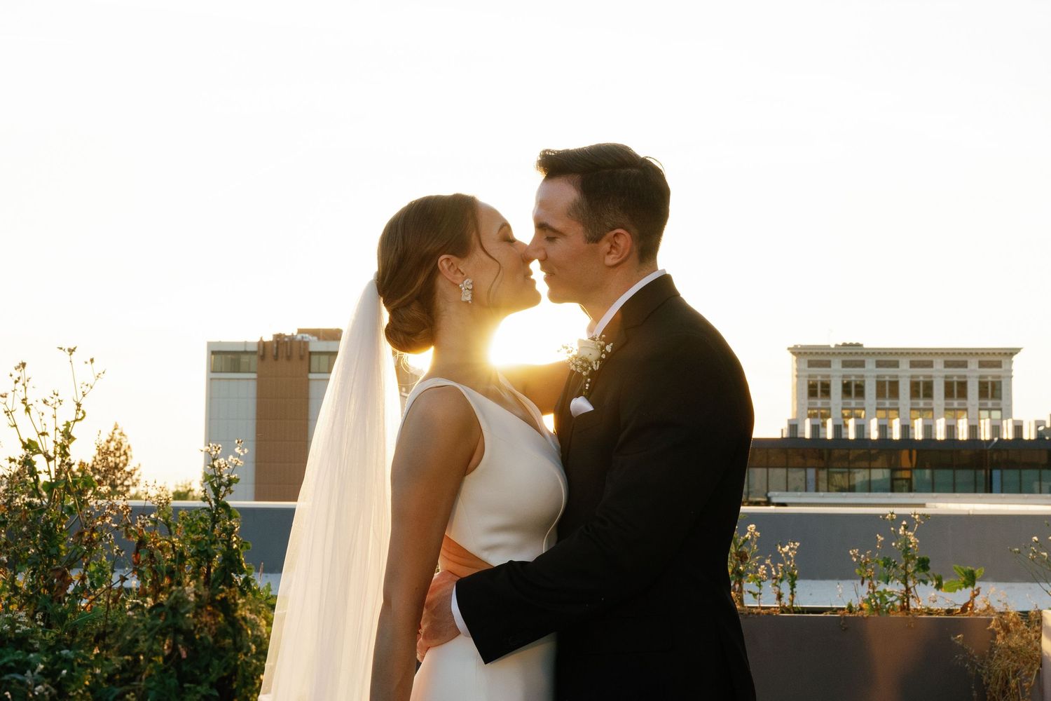 A bride and groom share a romantic kiss at sunset on a rooftop with golden sunlight glowing behind them.