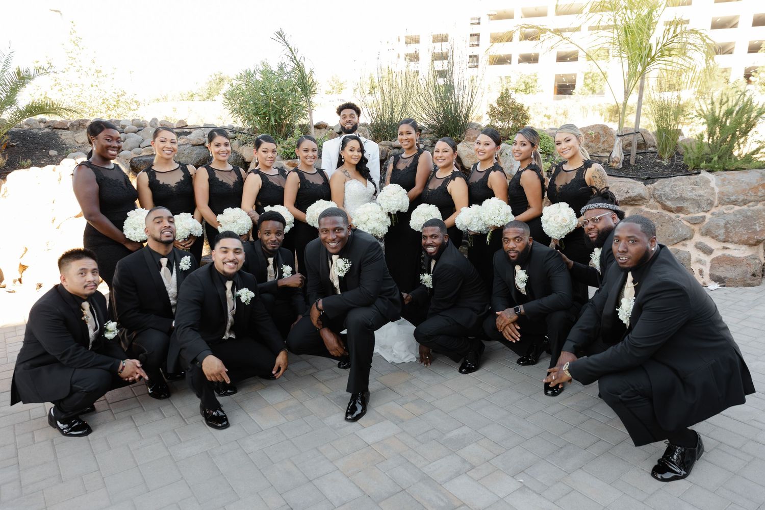 Groomsmen in black suits strike a fun pose lying on the ground in a curved formation at an outdoor wedding venue.
